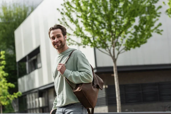 Smiling bearded man in sweatshirt holding leather strap of backpack near building outside — Stock Photo