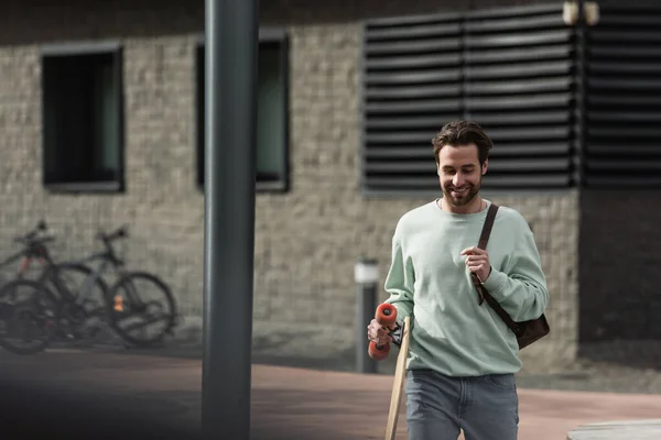 Sonriente hombre barbudo en sudadera sosteniendo longboard y correa de cuero de la mochila exterior - foto de stock