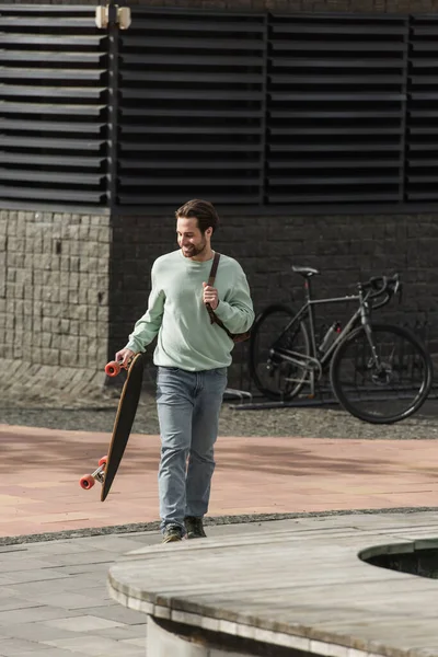 Full length of happy bearded man in sweatshirt holding longboard and leather strap of backpack while walking outside — Stock Photo