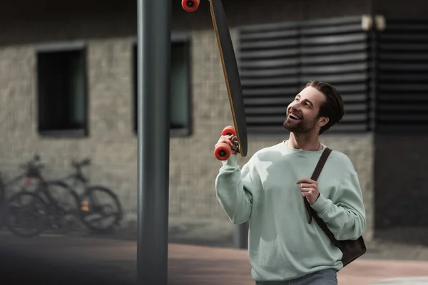 Cheerful bearded man in sweatshirt holding longboard and leather strap of backpack outside — Stock Photo