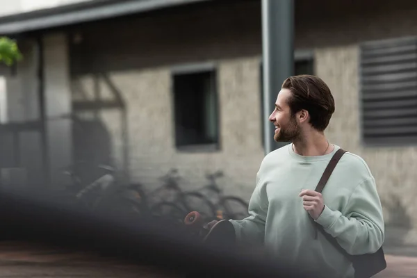 Happy bearded man in sweatshirt holding longboard and leather strap of backpack outside — Stock Photo