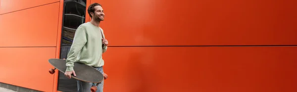 Hombre barbudo feliz en sudadera sosteniendo longboard cerca de la pared naranja, bandera - foto de stock