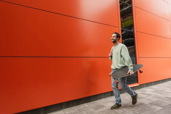 Hombre barbudo feliz en sudadera sosteniendo longboard y caminando cerca de la pared naranja - foto de stock