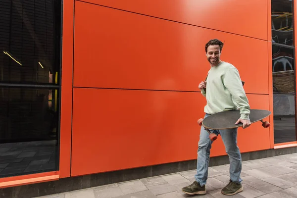 Cheerful bearded man in sweatshirt holding longboard while walking near orange wall — Stock Photo