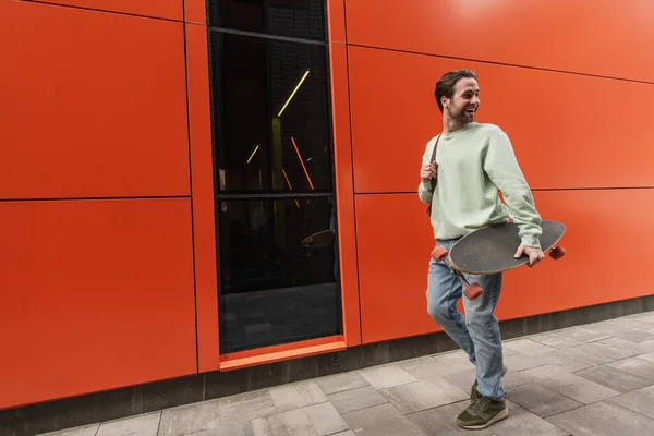 Allegro uomo in felpa tenendo longboard e camminando vicino alla parete arancione — Foto stock