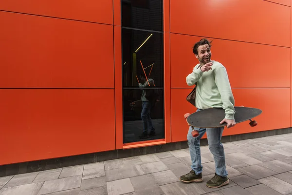 Smiling man in sweatshirt holding longboard while pointing away with finger near orange wall — Stock Photo