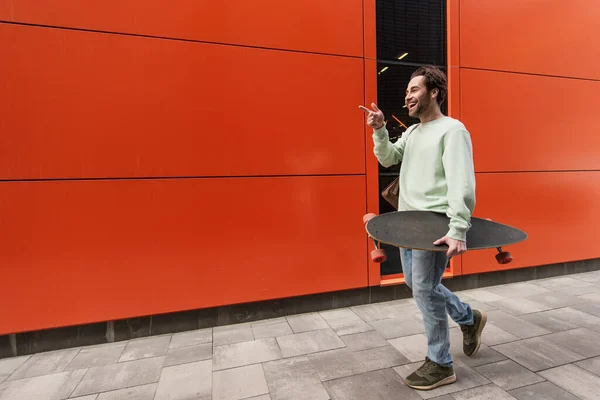 Sonriente hombre en sudadera sosteniendo longboard mientras señala con el dedo cerca de la pared naranja - foto de stock