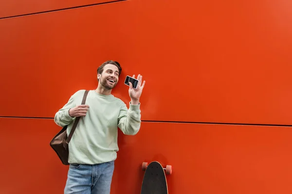 Cheerful man in sweatshirt holding smartphone with blank screen and waving hand near orange wall — Stock Photo