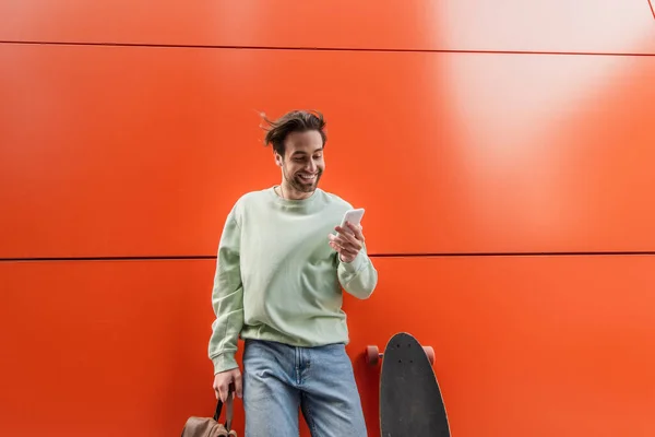 Hombre alegre en sudadera que sostiene la mochila y el uso de teléfono inteligente cerca de longboard y pared naranja - foto de stock