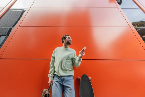 Low angle view of cheerful man in sweatshirt holding backpack and smartphone near longboard and orange wall — Stock Photo