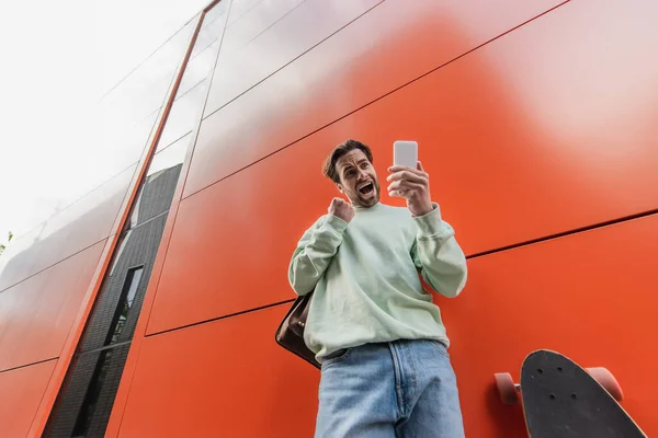 Visão de baixo ângulo do homem animado em camisola segurando smartphone perto de longboard e parede laranja — Fotografia de Stock