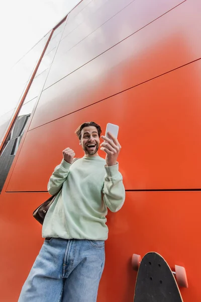 Low angle view of amazed man in sweatshirt holding smartphone near orange wall — Stock Photo