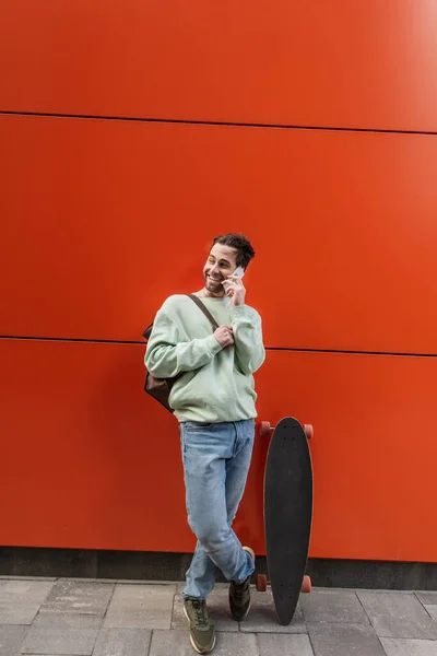 Hombre feliz en sudadera hablando en el teléfono celular mientras está de pie cerca de la pared naranja - foto de stock