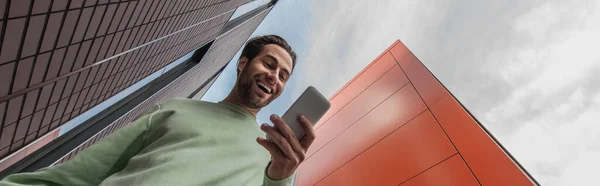 Vista de ángulo bajo del hombre sorprendido en la sudadera que sostiene el teléfono inteligente cerca de edificios, pancarta - foto de stock