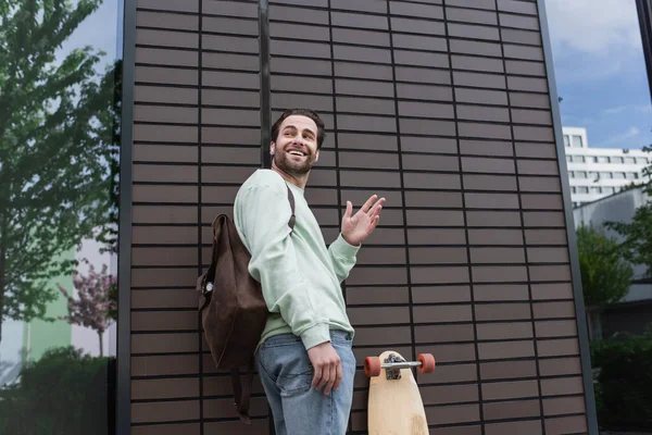 Glücklicher Mann in Sweatshirt und drahtlosen Kopfhörern, der draußen mit der Hand winkt — Stockfoto