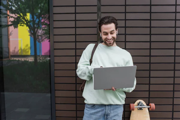 Bearded freelancer in sweatshirt and wireless earphones using laptop outside — Stock Photo