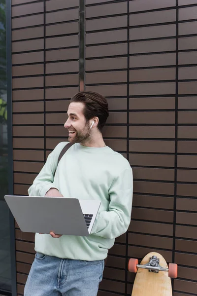 Feliz freelancer en sudadera y auriculares inalámbricos utilizando el ordenador portátil fuera - foto de stock