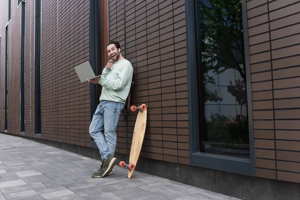 Sorprendido freelancer en sudadera y auriculares inalámbricos sosteniendo portátil y señalando con el dedo fuera - foto de stock