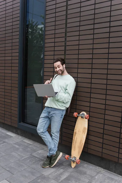 Full length of happy freelancer in sweatshirt and wireless earphones using laptop outside — Stock Photo