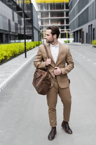 Full length of bearded businessman in suit holding leather strap of bag outside — Stock Photo