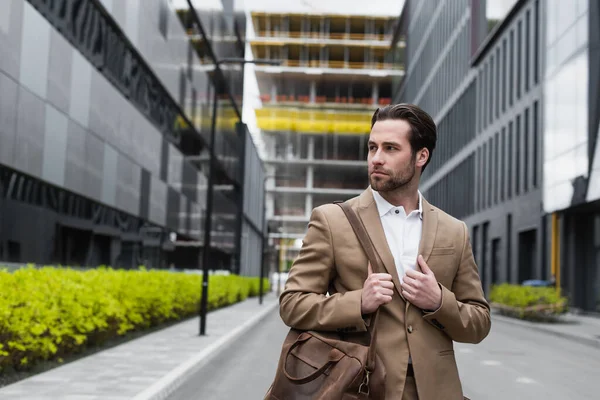 Young businessman in suit holding leather strap of bag on urban street — Stock Photo