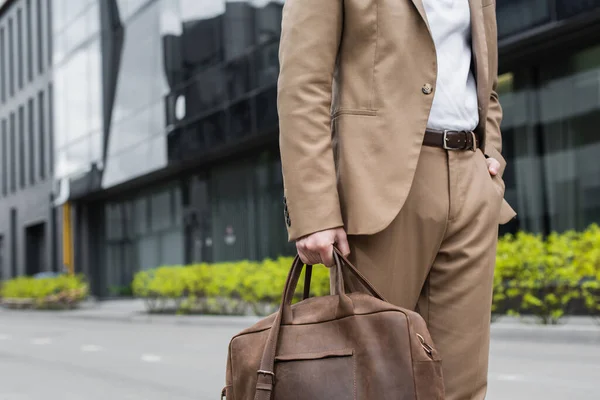 Cropped view of businessman in suit holding leather bag and standing with hand in pocket on urban street — Stock Photo