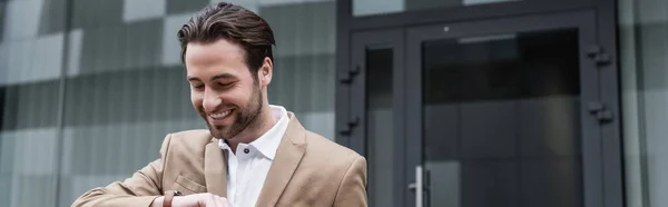 Hombre de negocios barbudo feliz en traje mirando reloj de pulsera, bandera - foto de stock