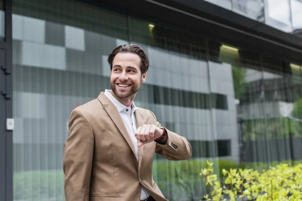 Hombre de negocios barbudo feliz en desgaste formal con reloj de pulsera mirando hacia otro lado - foto de stock