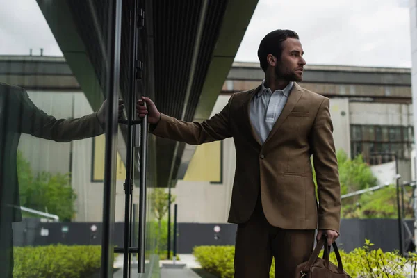 Hombre de negocios barbudo en ropa formal que sostiene el bolso de cuero cerca del edificio exterior - foto de stock