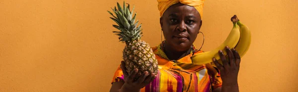 African american woman in yellow turban and colorful dress holding pineapple and bananas on orange, banner — Stock Photo