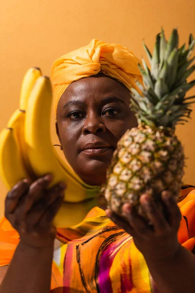 Mujer afroamericana en turbante amarillo y vestido colorido sosteniendo piña y plátanos en naranja - foto de stock