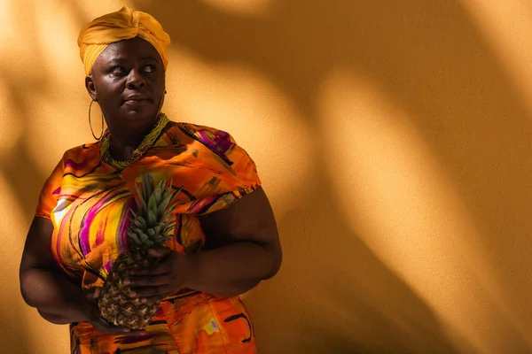 Middle aged african american woman in yellow turban and colorful dress holding pineapple on orange — Stock Photo