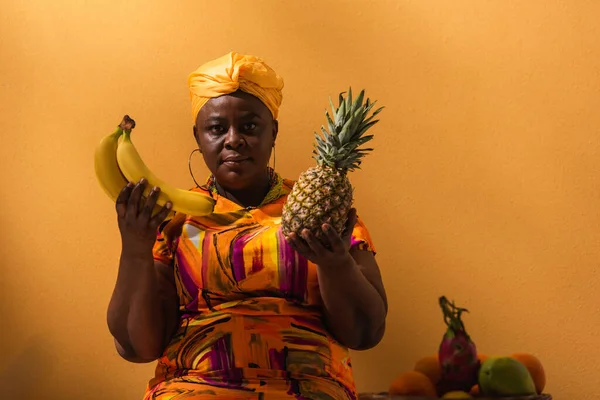 Afrikanische Amerikanerin mittleren Alters hält Ananas und Bananen in der Nähe von Früchten auf Orange — Stockfoto