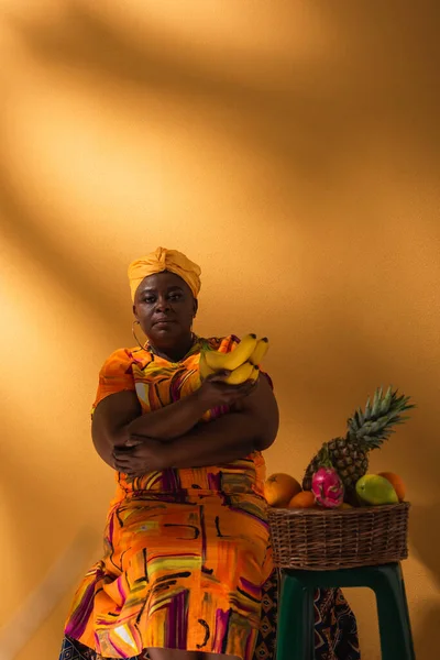 Mulher afro-americana de meia idade sentada perto de frutas e segurando bananas em laranja — Fotografia de Stock