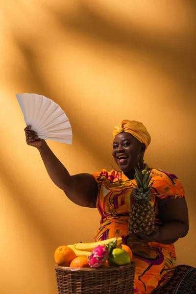 Lächelnde Afroamerikanerin mittleren Alters, die Ananas in der Nähe von Früchten hält und mit Fächer auf Orange wedelt — Stockfoto