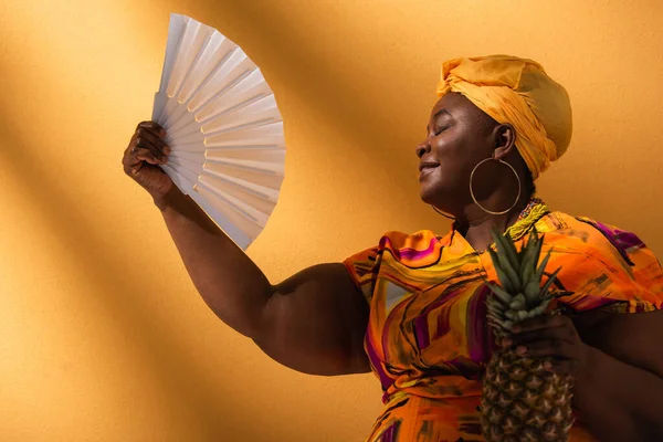 Middle aged african american woman sitting with closed eyes, holding pineapple and waving with fan on orange — Stock Photo