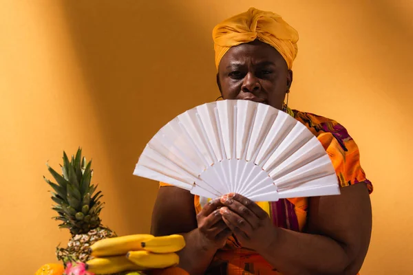 Seria de mediana edad afroamericana mujer sentada cerca de frutas y sosteniendo ventilador en naranja - foto de stock