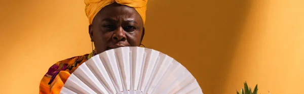 Mujer afroamericana de mediana edad serio que cubre la cara con ventilador en naranja, bandera - foto de stock