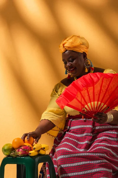 Fröhliche afrikanisch-amerikanische Frau mittleren Alters berührt Früchte und hält Fächer auf gelb — Stockfoto