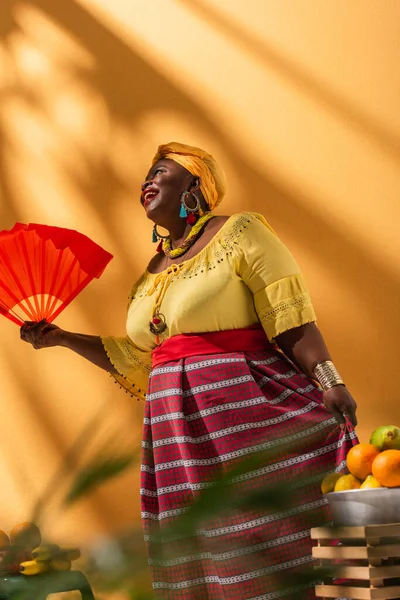 Vista a basso angolo di positiva donna afro-americana di mezza età che vende frutta e tiene ventilatore su arancione — Foto stock