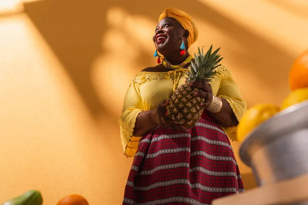 Low angle view of smiling middle aged african american woman selling fruits with pineapple in hand on orange — Stock Photo