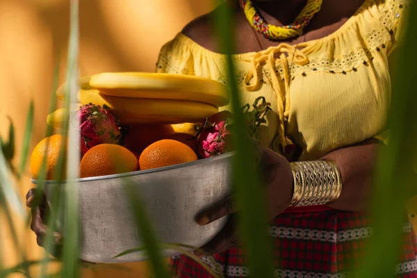 Vista recortada de mujer afroamericana de mediana edad en blusa amarilla sosteniendo un tazón de metal con frutas en naranja - foto de stock