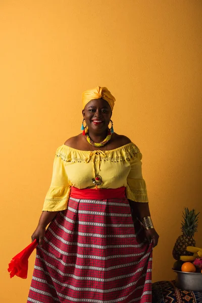 Donna afroamericana di mezza età in abiti luminosi e turbante in piedi vicino a frutti in ciotola di metallo su giallo — Foto stock