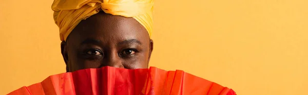 Portrait of middle aged african american woman covering face with red fan on yellow, banner — Stock Photo
