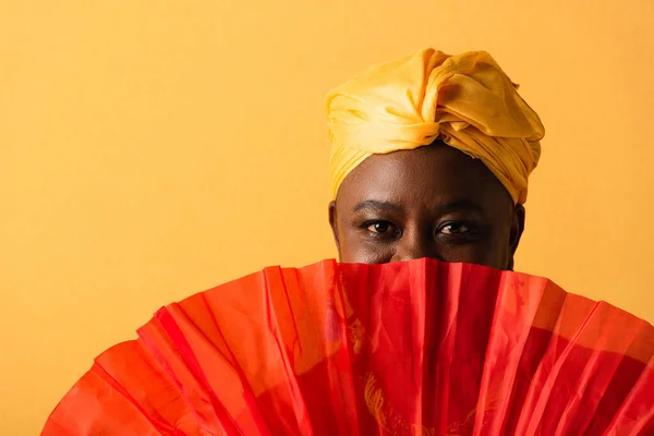Портрет афроамериканки середнього віку, що прикриває обличчя червоним вентилятором на жовтому — стокове фото