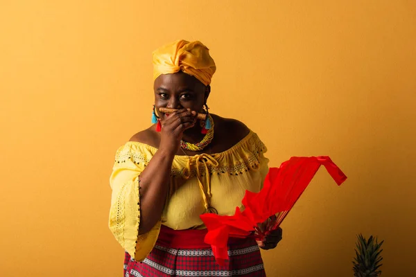 Middle aged african american woman in yellow turban and bright clothes smelling aroma of cigar on yellow — Stock Photo