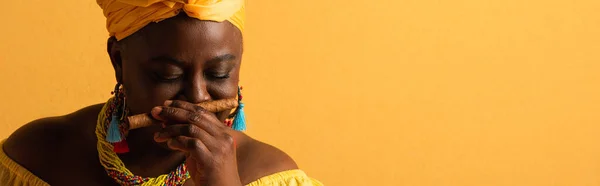 Middle aged african american woman in yellow turban smelling aroma of cigar with closed eyes on yellow, banner — Stock Photo