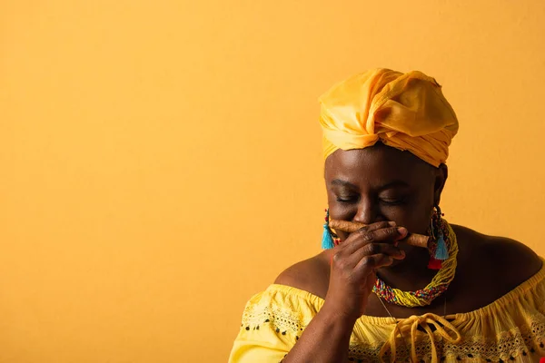 Middle aged african american woman in yellow turban smelling aroma of cigar with closed eyes on yellow — Stock Photo