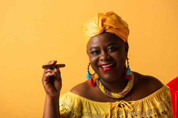 Sonriente mujer afroamericana de mediana edad en turbante amarillo y blusa sosteniendo cigarro en amarillo - foto de stock