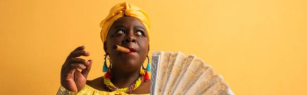 Middle aged african american woman in yellow turban and blouse holding fan and smoking cigar on yellow, banner — Stock Photo